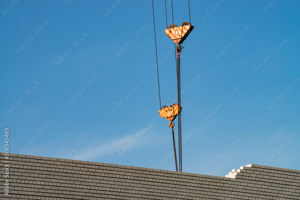 Hook of tower crane above unfinished building in construction site. Background image of white brick wall under blue sky. Background image of process building. Hook of tower crane above unfinished building in construction site. Background image of white brick wall under blue sky. Background image of process building.