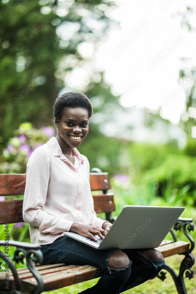 Young beauty afro american woman sitting on wooden bench in the park and working on laptop Young beauty afro american woman sitting on wooden bench in the park and working on laptop