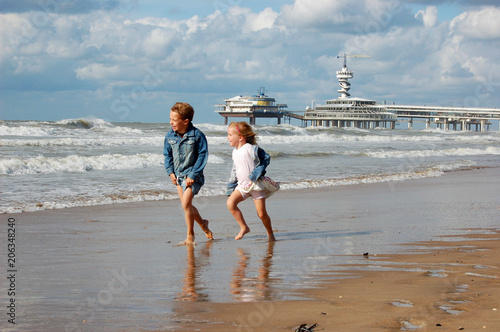 Little boy and girl running in denim clothing in the surf of the sea, with a pedestrian promenade in the background. Scheveningen, The Netherlands, Europe.
