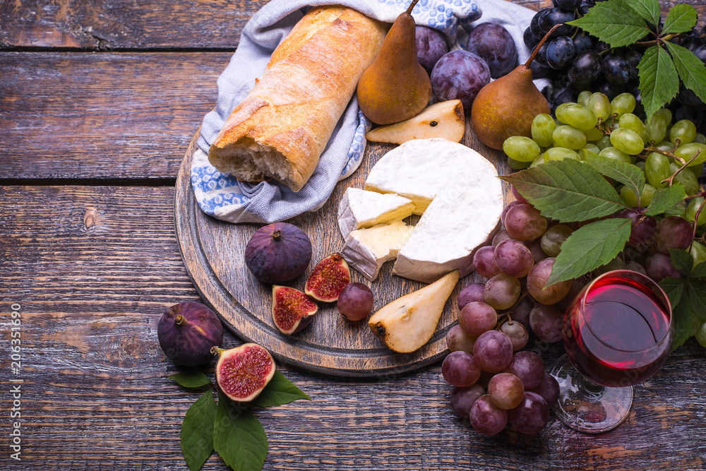 A glass of red wine, grapes, cheese, cork, corkscrew, white bread on a dark background