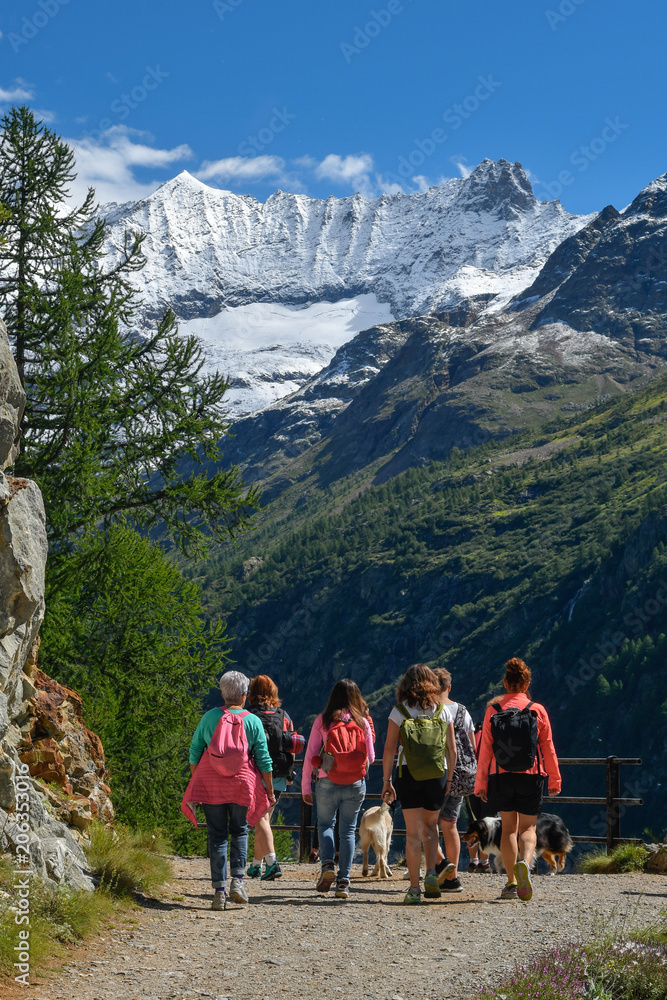 Group of people with backpacks walking on a mountain path along a turquoise lake with snowy peaks on background. A beautiful sunny day to trekking with family and friends climbing to the snow-covered 