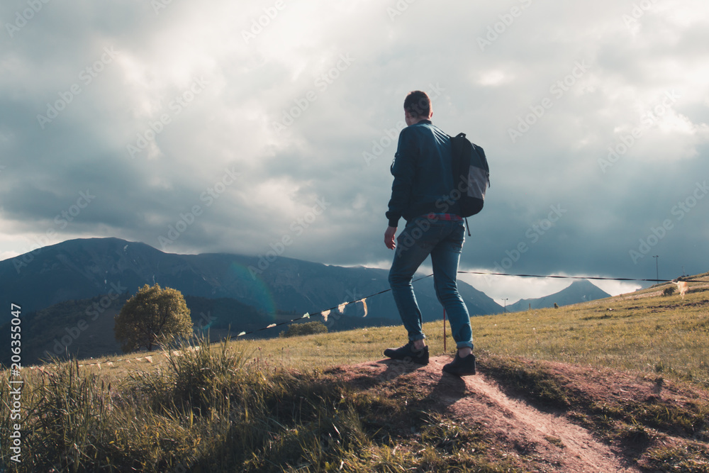 Lonely young tourist guy with backpack climbed to the top and looks at the beautiful mountain landscape before the storm Lonely young tourist guy with backpack climbed to the top and looks at the beautiful mountain landscape before the storm