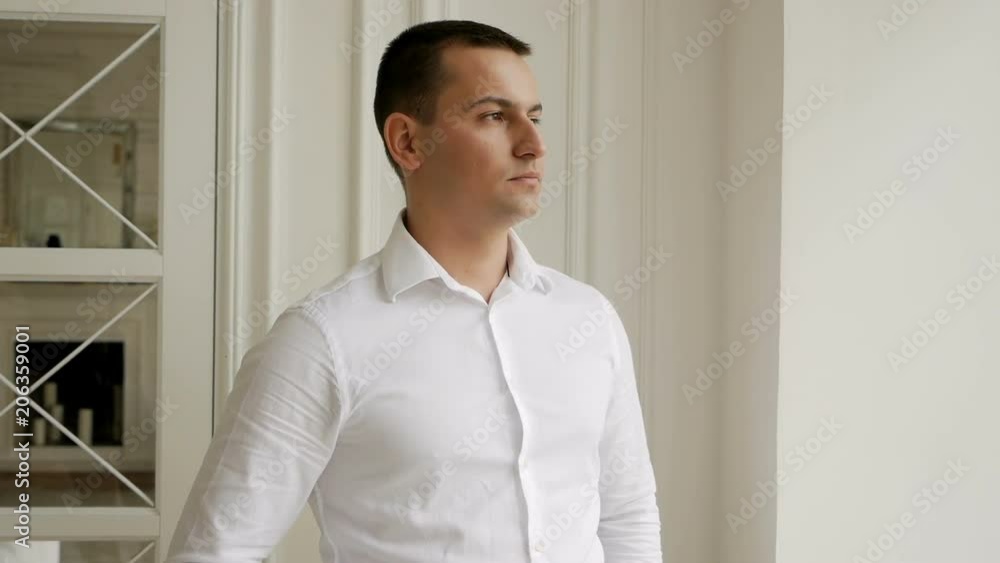 young man businessman in a white shirt stands at the window and turns his head towards the viewer young man businessman in a white shirt stands at the window and turns his head towards the viewer