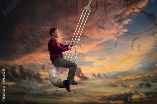 Man in red sweater sitting on the pendulum of a grandfather clock which is swinging in the air on twilight background photo