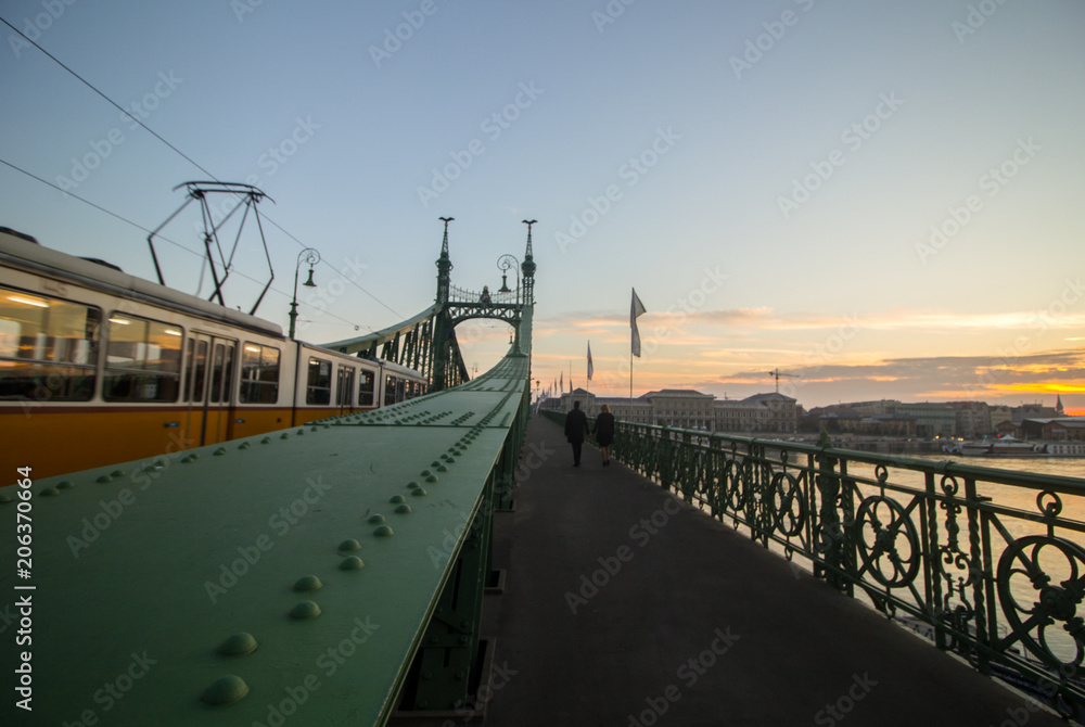 Couple in love is standing on the liberty bridge over the river Danube in Budapest. Sunrise in the big city. Dark silhouettes of man and woman walking in urban landscape. Yellow old tram going by.