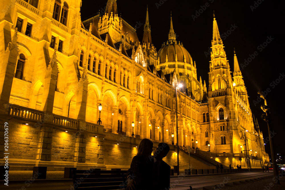 Couple in love is kissing and hugging near the night Budapest Parliament in yellow lights. European city travelling lifestyle photo. Man and woman silhouettes on the bench.