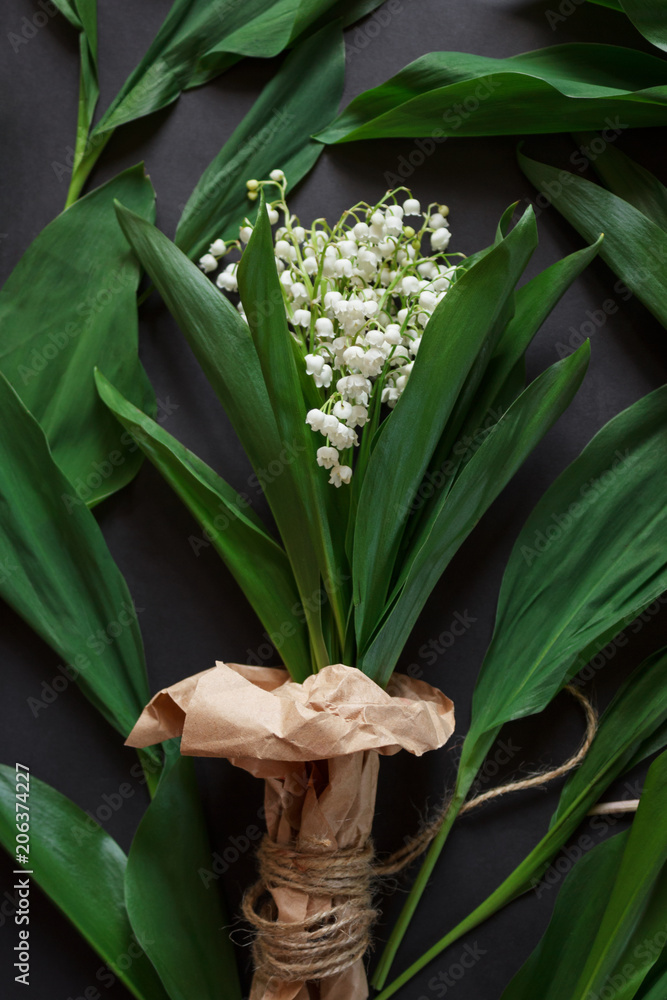 Obraz premium A bouquet of lilies of the valley in kraft paper on a dark background with a pattern of green leaves. Flat lay.