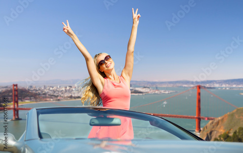 travel, summer holidays, road trip and people concept - happy young woman wearing sunglasses in convertible car showing peace hand sign over golden gate bridge in san francisco bay background