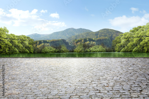 A square made of stone cubes against the background of a green park with a lake and mountains in the background.