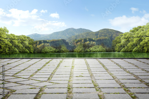 Empty square made of stone cubes against the background of a green park with a lake and mountains and sky in the background.