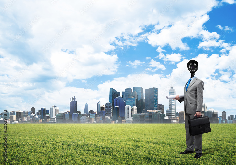 Camera headed man standing on green grass against modern cityscape Camera headed man standing on green grass against modern cityscape