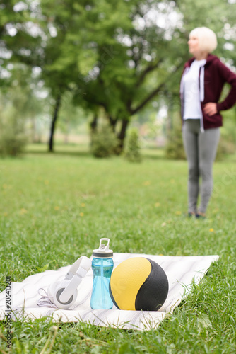 selective focus of woman in park with medicine ball, water and headphones on yoga mat on foreground