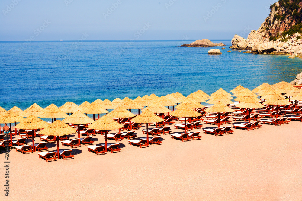 Obraz premium Beach Umbrella made of palm leafs on the background of an exotic beach in Montenegro. Relaxing area: sun beds in the shadow of rustic umbrella. Sea view