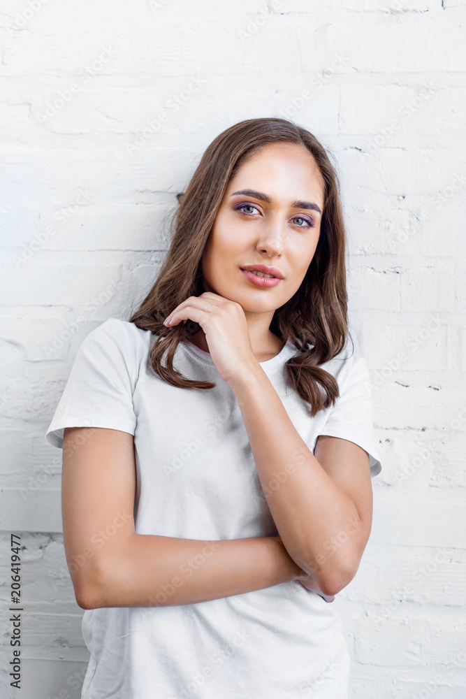beautiful young woman in white t-shirt standing near white brick wall and looking at camera