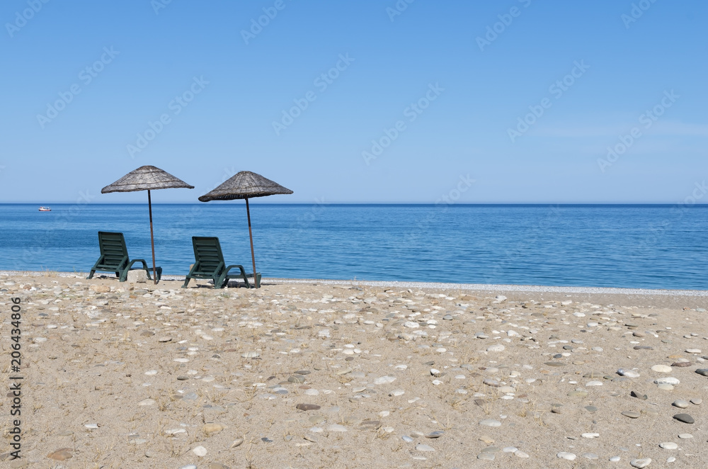 Beach chairs under thatched umbrellas with blue sea and sky on sunny summer day. A beautiful place to relax. Sunbeds on the beach against the azure or turquoise calm sea with boat Beach chairs under thatched umbrellas with blue sea and sky on sunny summer day. A beautiful place to relax. Sunbeds on the beach against the azure or turquoise calm sea with boat