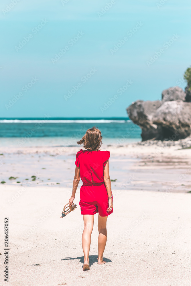 Girl in a red dress walking barefoot on the sea shore. Tropical beach, Bali island. Sunny day.