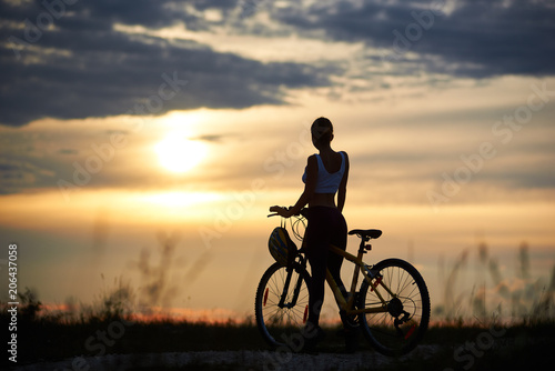 Silhouette of sporty female cyclist, wearing sportswear, standing near her bicycle on trail. Slender incognito woman enjoying nature and observing wonderful landscapes and amazing sunset.