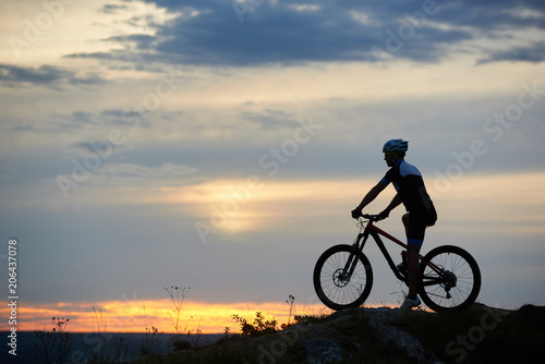 Sport and recreational activities at fresh air under evening sky. Incognito cyclist sitting on bicycle and riding bike in twilight. Active man posing and wearing sportswear and helmet.