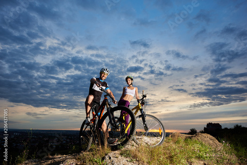 Smiling young couple of cyclists in helmets sitting on mountain bicycles and looking at camera. Athletic boyfriend and beautiful girlfriend posing on rock hill against cloudy evening sky background.