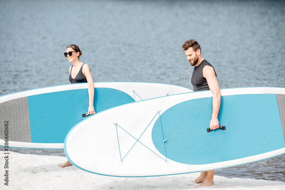 Φωτογραφία Beautiful couple in black swimwear walking with standup paddleboard on the beach
