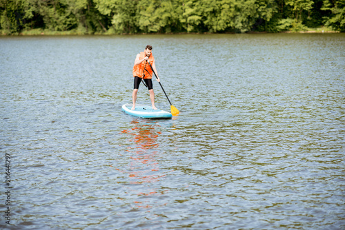 Man in life vest swimming on the standup paddleboard on the lake near the green forest
