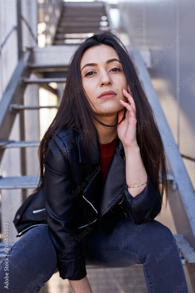 Fototapeta premium Young stylish woman in jeans and leather jacket resting on the park stairs. Attractive girl with long hair sitting on steps