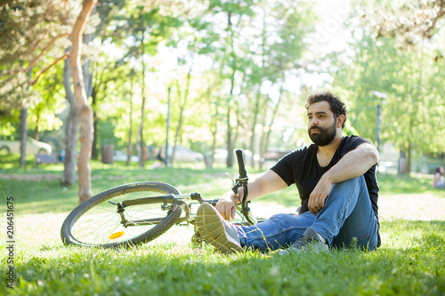 Young bearded man resting on the grass in the park next to his bicycle