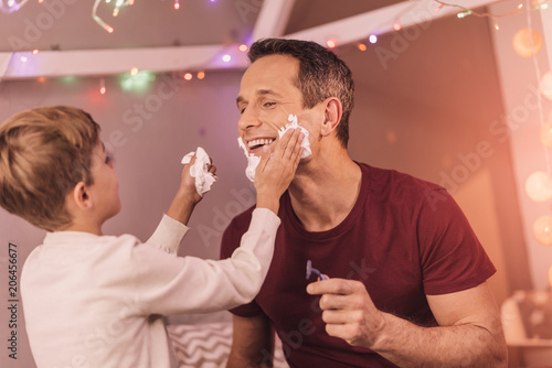 Positive mood. Delighted joyful nice man smiling to his son and preparing to shave while being at home