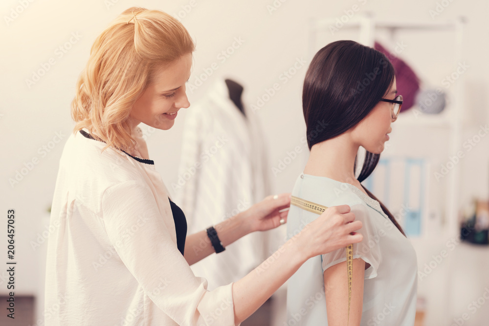 Delicate figure. Close up of young skilled designer taking measurements of beautiful girl while standing behind her back Delicate figure. Close up of young skilled designer taking measurements of beautiful girl while standing behind her back