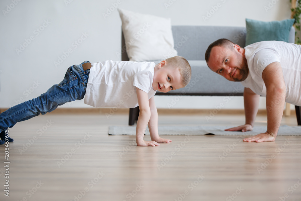 Photo of young father and son pushing up on floor against sofa Photo of young father and son pushing up on floor against sofa