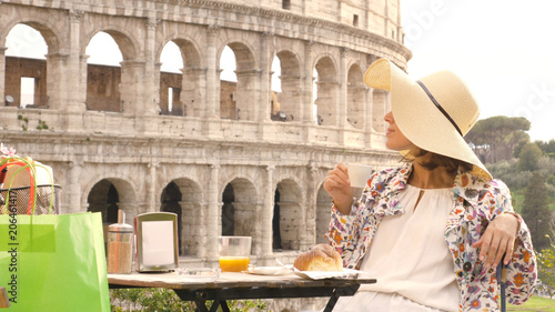 Photography Happy young woman tourist drinking coffee and juice with cornetto at the table outside a bar restaurant in front of the Colosseum in Rome
