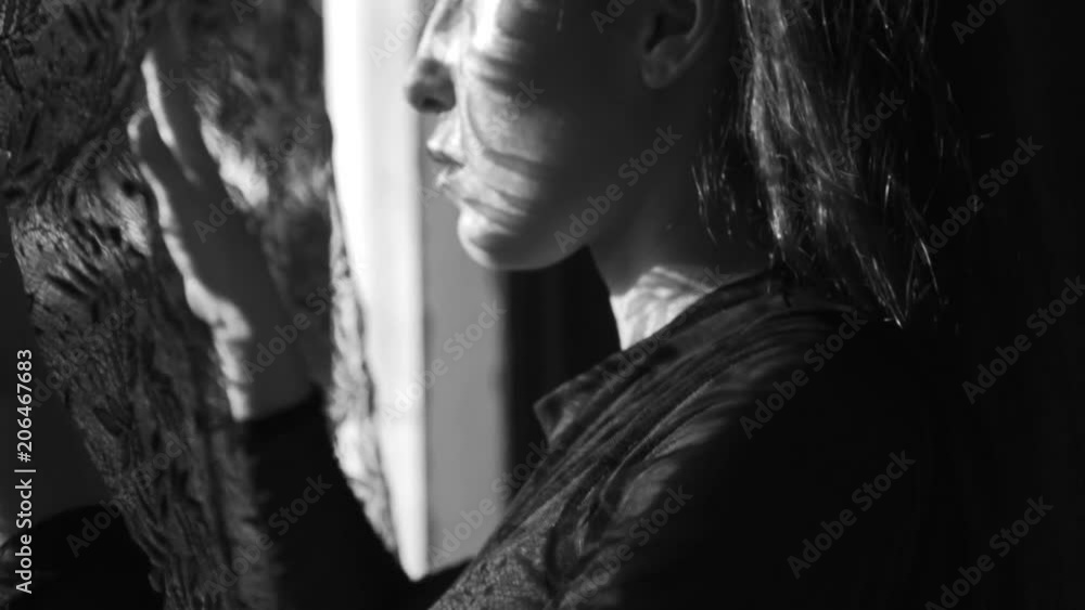 Beautiful woman looking through the window, she has got lace curtain pattern shadow and sun rays on her face - black and white video in slow motion Beautiful woman looking through the window, she has got lace curtain pattern shadow and sun rays on her face - black and white video in slow motion