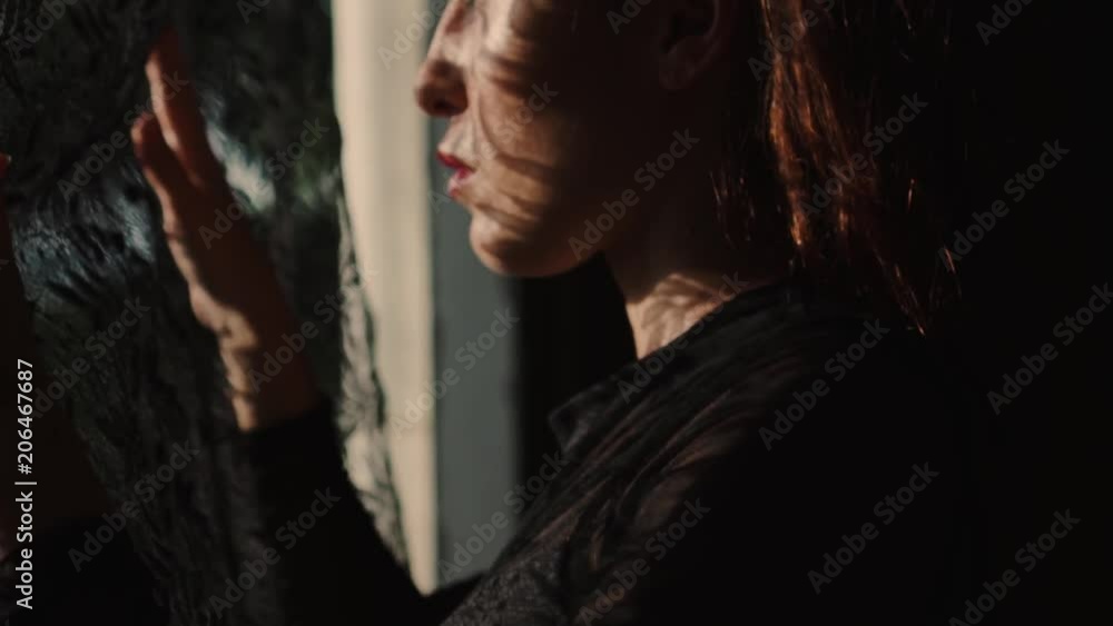 Beautiful woman looking through the window, she has got lace curtain pattern shadow and sun rays on her face - video in slow motion Beautiful woman looking through the window, she has got lace curtain pattern shadow and sun rays on her face - video in slow motion