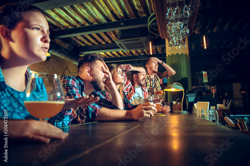 Upset male and female friends watching sport game or football match and drinking beer at bar or pub.