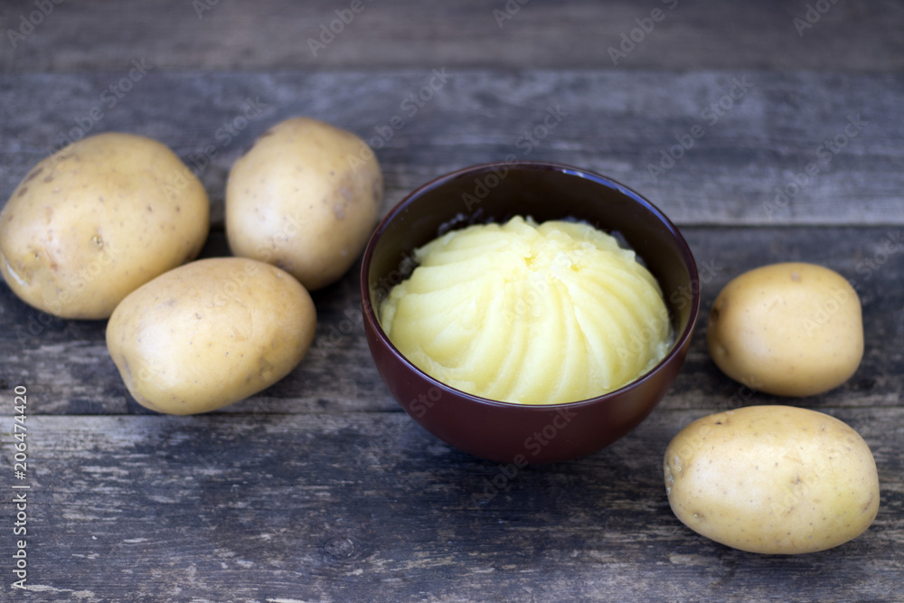 Mashed potatoes and raw potatoes side by side, located on a wooden background, rustic style close-up