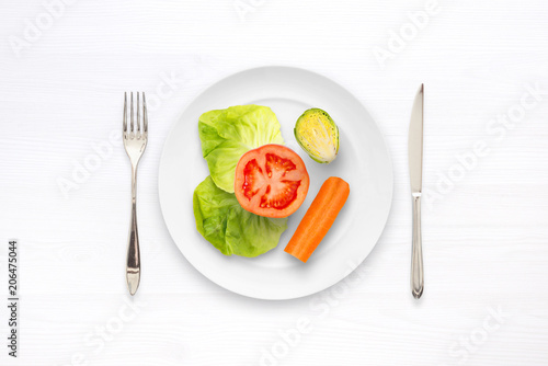 Diet meal with vegetables. In the plate are salad, tomato, cucumber and carrots. Next to the plate are a spoon and a fork. Flat view, white space.