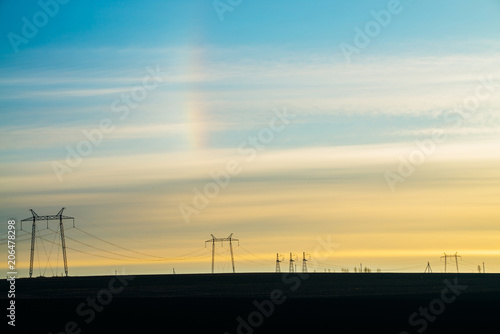 Power lines on background of sunny sky with rainbow close-up. Silhouette of electric pole with copy space. Wires of high voltage above ground. Electricity industry.