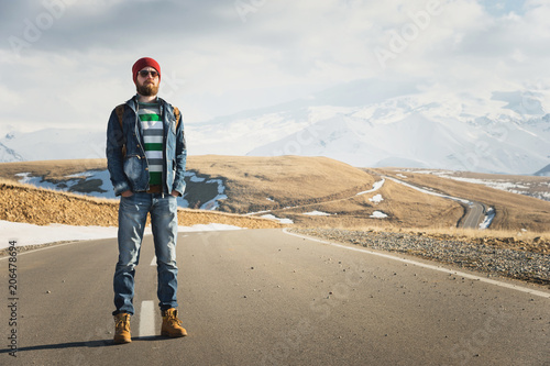 A stylish bearded hipster in sunglasses with a vintage backpack stands on a country road asphalt on a sunny day. The concept of hitchhiking and hiking © yanik88