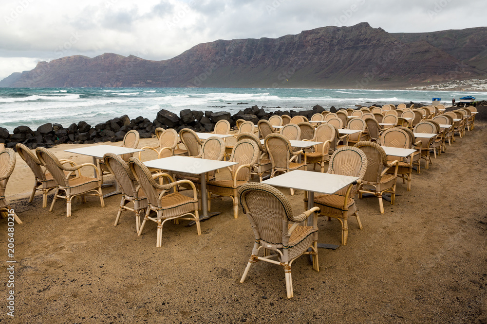 LANZAROTE, CANARY ISLANDS, SPAIN: Low season - empty tables and chairs on the terraces of Famara, with the sea and cliffs in the background. LANZAROTE, CANARY ISLANDS, SPAIN: Low season - empty tables and chairs on the terraces of Famara, with the sea and cliffs in the background.