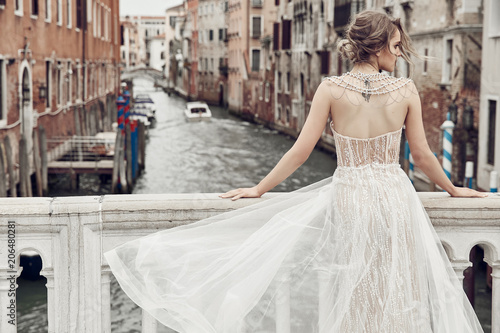 Beautiful bride with tattoo on back, dressed in elegant, luxury ivory dress and updo hairstyle, posing on a bridge over the canal in Venice