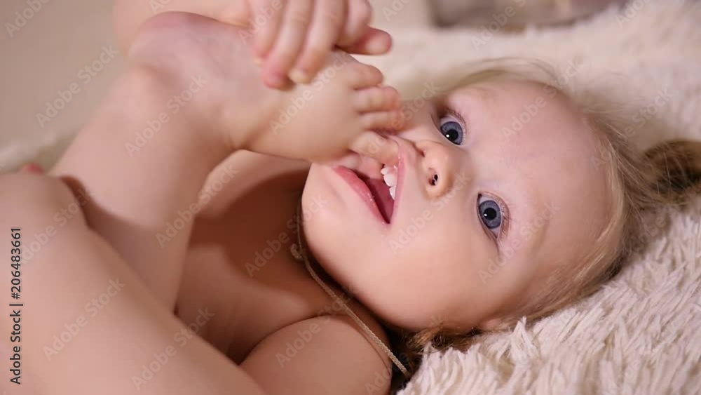 Little cute toddler baby girl lie on bed bite and suck her foot toes playing with leg Little cute toddler baby girl lie on bed bite and suck her foot toes playing with leg