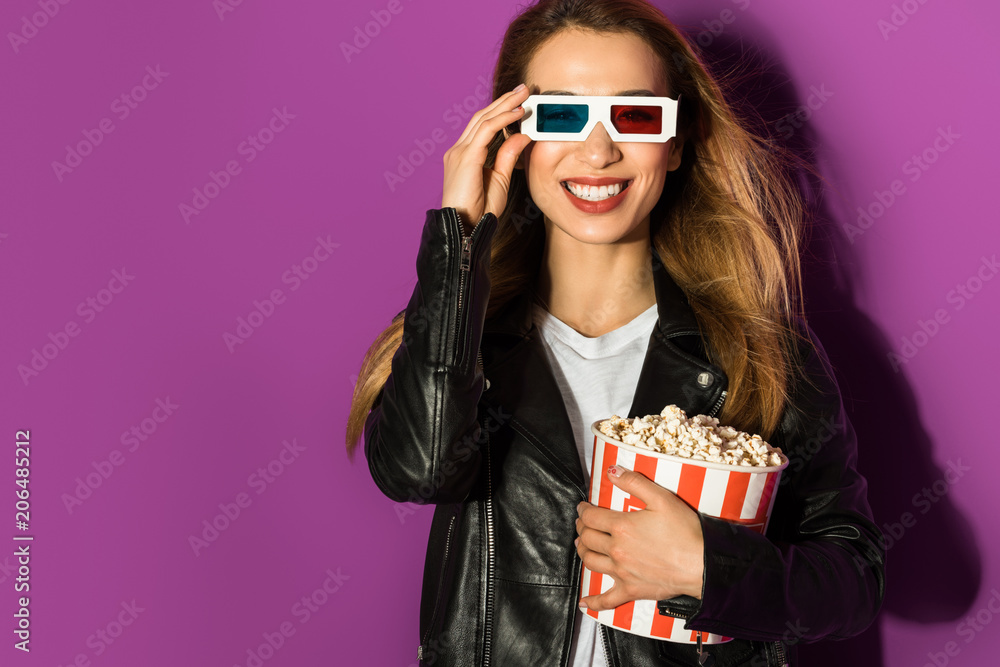 Obraz premium beautiful young woman in 3d glasses holding box of popcorn and smiling at camera isolated on violet