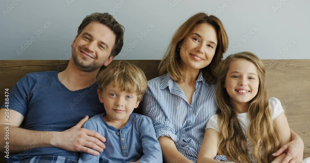 Portrait shot of the good cheerful Caucasian parents hugging their small son and daughter while sitting together and posing to the camera in the bed in the morning. Indoors Portrait shot of the good cheerful Caucasian parents hugging their small son and daughter while sitting together and posing to the camera in the bed in the morning. Indoors