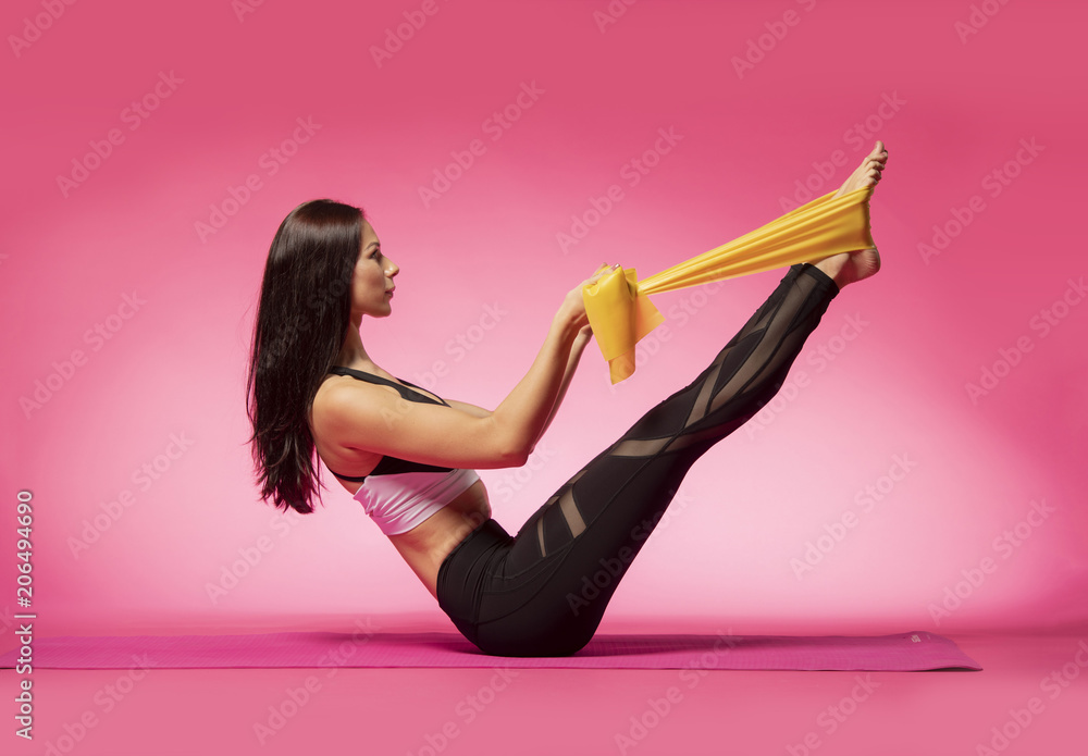 Long haired beautiful pilates or yoga athlete does a graceful pose with a  yellow rubber band while wearing a tight sports outfit against a pink  background in a studio Stock Photo