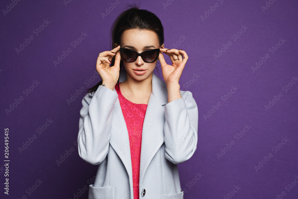 Beautiful and fashionable brunette model girl in red dress and coat adjusting her sunglasses and posing at violet background Beautiful and fashionable brunette model girl in red dress and coat adjusting her sunglasses and posing at violet background