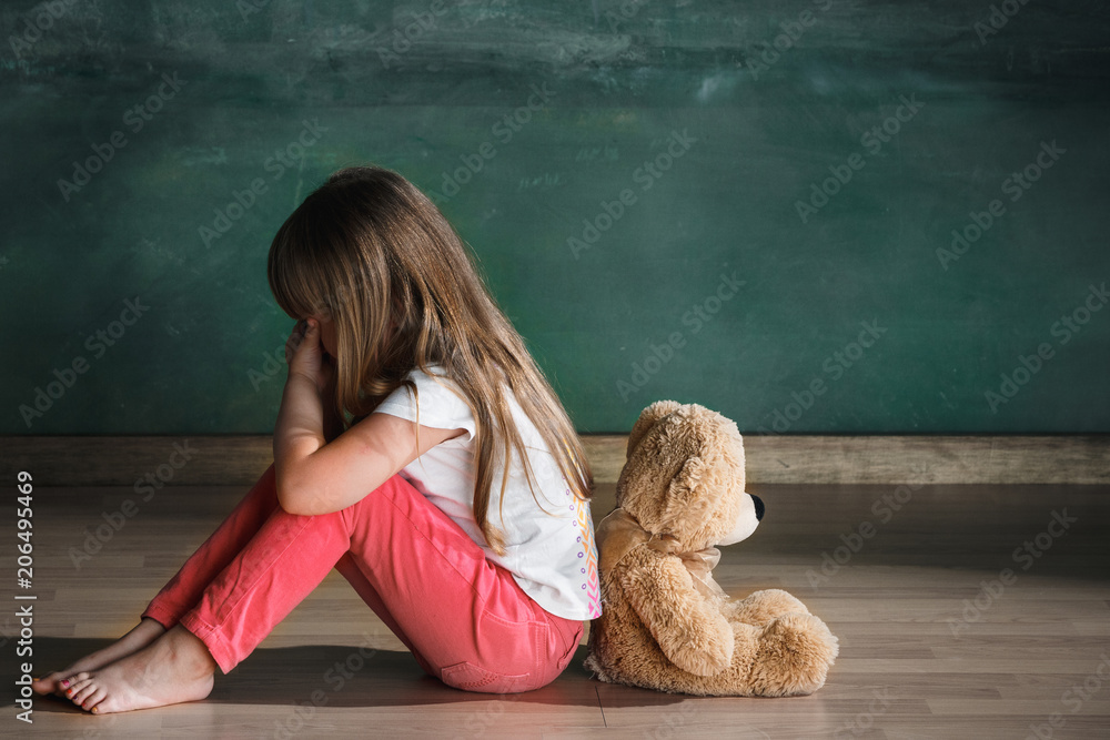 Little girl with teddy bear sitting on floor in empty room. Autism concept - obrazy, fototapety, plakaty 