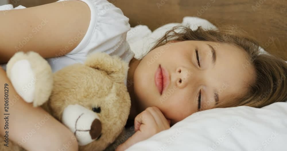 Close up of the cute small blond girl with a teddy bear in hands sleeping sweet in a bed in the morning. Inside Close up of the cute small blond girl with a teddy bear in hands sleeping sweet in a bed in the morning. Inside