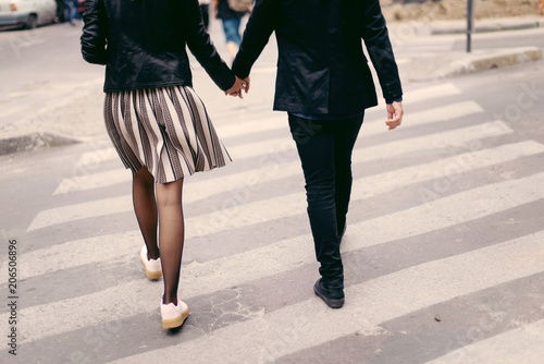 Young fashion couple holding hands and crossing the road. Close up man and woman legs on the crosswalk. Outdoors in the street. Back view shot