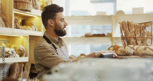 Young handsome man vendor in the apron bringing fresh bread to the counter in the bakery shop. Indoors