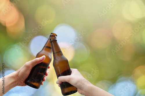 Hands holding two beer bottle and happy enjoying harvest time together to clinking glasses at outdoor party on beautiful bokeh light background.Celebration drinking beer in pub,bar.                  